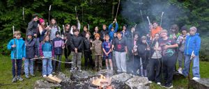 Eine Gruppe von Kindern steht um ein Lagerfeuer im Wald und posiert für ein Foto. Sie tragen Winterkleidung und sind von Bäumen umgeben.