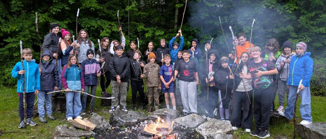 Eine Gruppe von Kindern steht um ein Lagerfeuer im Wald und posiert für ein Foto. Sie tragen Winterkleidung und sind von Bäumen umgeben.