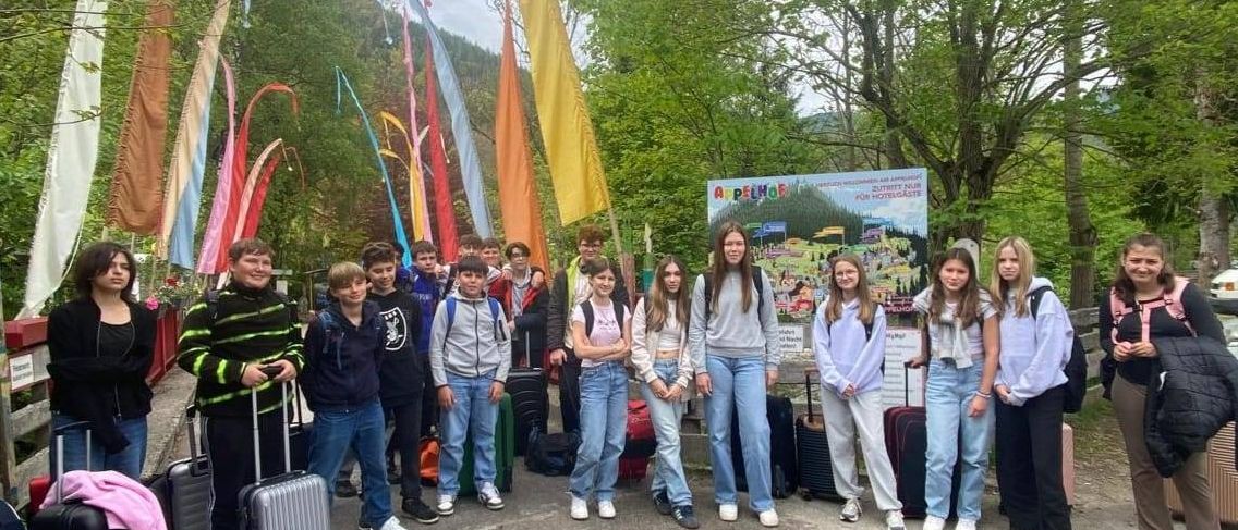 Eine Gruppe von Schülern steht mit Gepäck, bunten Fahnen und einer malerischen Kulisse auf einer Straße. Dahinter sind Bäume und Berge zu sehen. Ein Schild im Hintergrund lautet Appelhof.