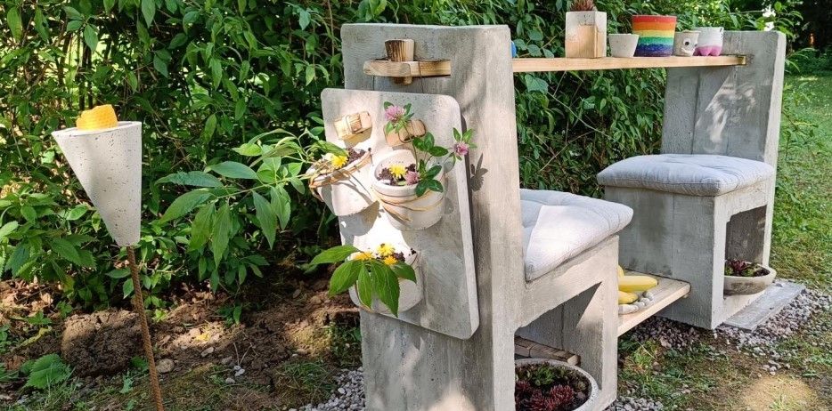 Eine Betonbank mit Topfpflanzen und einem Holztablett, in einem Garten mit üppigem grünem Laub aufgestellt.