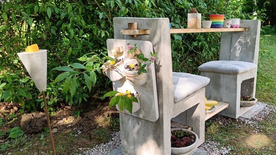 Eine Betonbank mit Topfpflanzen und einem Holztablett, in einem Garten mit üppigem grünem Laub aufgestellt.