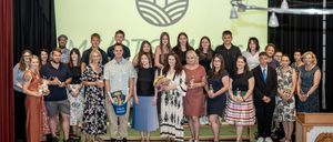 Eine Gruppe Erwachsener steht mit Blumen, Flaschen und einem blauen Eimer auf der Bühne. Sie scheinen ein Ereignis zu feiern.