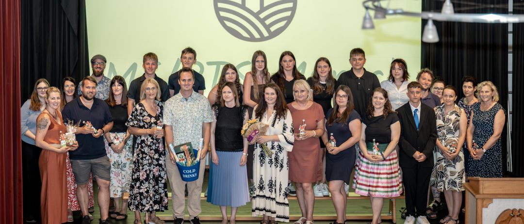 Eine Gruppe Erwachsener steht mit Blumen, Flaschen und einem blauen Eimer auf der Bühne. Sie scheinen ein Ereignis zu feiern.