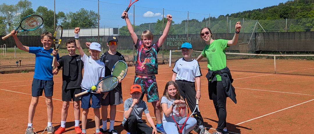 Eine Gruppe von Kindern und zwei Erwachsenen posiert auf einem Tennisplatz. Sie halten alle Tennisschläger in den Händen und scheinen für ein Foto zu lächeln.