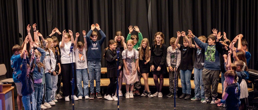 Eine Gruppe von Kindern in Freizeitkleidung steht mit Mikrofonen auf der Bühne und tritt vor einem schwarzen Vorhang auf. Einige von ihnen heben die Hände.