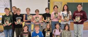 Eine Gruppe junger Schüler, sowohl Jungen als auch Mädchen, halten Bücher mit einem Charakter auf dem Cover hoch. Sie sind in einem Klassenzimmer mit einer Tafel, einer schwarzen Tafel und gelben Wänden.