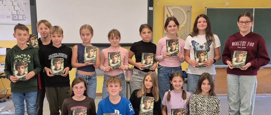 Eine Gruppe junger Schüler, sowohl Jungen als auch Mädchen, halten Bücher mit einem Charakter auf dem Cover hoch. Sie sind in einem Klassenzimmer mit einer Tafel, einer schwarzen Tafel und gelben Wänden.