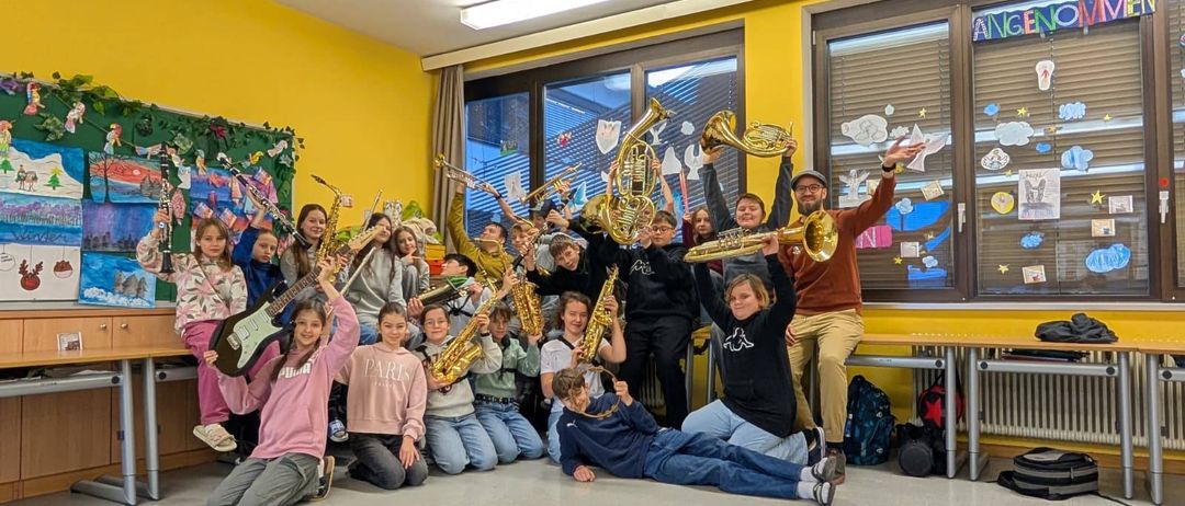 Eine Gruppe junger Musiker posiert für ein Foto in einem gelb gestrichenen Raum und hält Blech- und Saxophoninstrumente. Sie sitzen und stehen, mit zwei Erwachsenen in der Nähe. Der Raum hat Fenster mit Jalousien und eine Deckenleuchte.