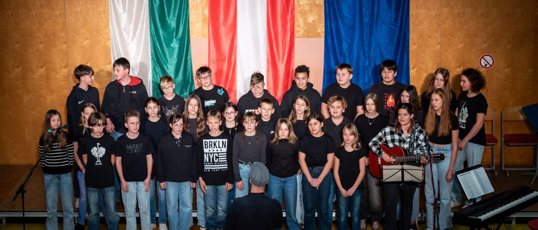 Eine Gruppe junger Menschen steht vor drei Flaggen, wahrscheinlich für eine Choraufführung. Die Flaggen sind grün, rot und weiß sowie blau mit Sternen. Eine Frau spielt Gitarre und ein Mann steht davor.