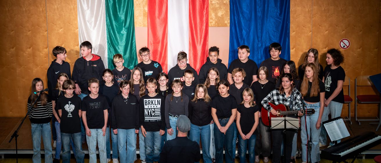 Eine Gruppe junger Menschen steht vor drei Flaggen, wahrscheinlich für eine Choraufführung. Die Flaggen sind grün, rot und weiß sowie blau mit Sternen. Eine Frau spielt Gitarre und ein Mann steht davor.