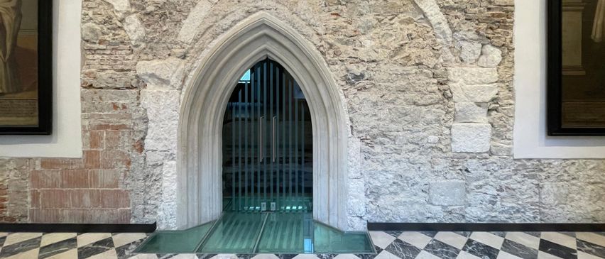 Ein steinerner Torbogen mit einer Glastür führt zu einem Raum mit einem gemusterten Boden.