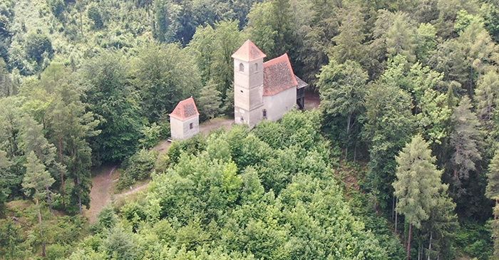 Ein Luftbild einer kleinen Kirche, eingebettet in einen dichten Wald, mit einem Turm und einem kleineren Gebäude in der Nähe.