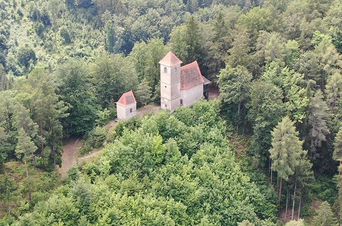 Ein Luftbild eines kleinen Gebäudes mit einem Turm, möglicherweise eine Kapelle, auf einem Hang inmitten eines dichten Waldes.
