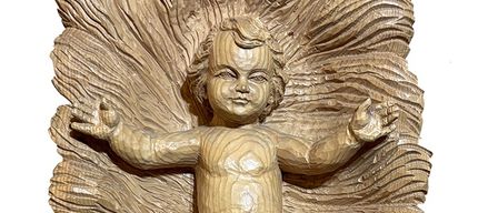 Eine Holzskulptur eines Babys, sitzend auf einer gebogenen, strukturierten Oberfläche, mit einem kleinen Gewand, einem ruhigen Gesichtsausdruck und offenen Armen.