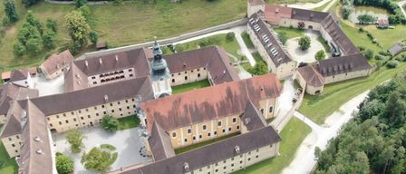 Eine Luftaufnahme eines großen historischen Gebäudes mit mehreren Flügeln, braunen Dächern und vielen Fenstern. Das Gebäude ist von grünen Rasenflächen und Bäumen umgeben, mit einem Turm in der Mitte.