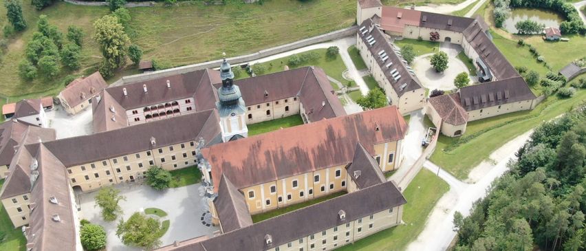 Eine Luftaufnahme eines großen historischen Gebäudes mit mehreren Flügeln, braunen Dächern und vielen Fenstern. Das Gebäude ist von grünen Rasenflächen und Bäumen umgeben, mit einem Turm in der Mitte.