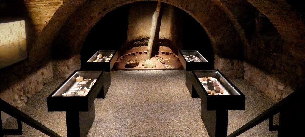 Ein schwach beleuchteter Untergrundraum mit Steinmauern und gewölbten Öffnungen. Drei schwarze Vitrinen mit Artefakten sind vor einem zentralen Steinpfeiler angeordnet.