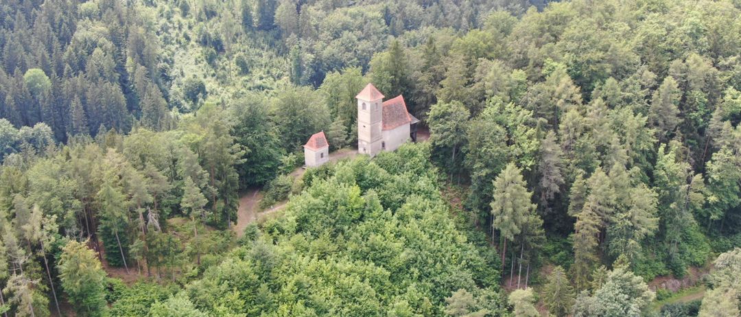 Eine Luftaufnahme einer kleinen Kapelle, umgeben von dichtem Wald mit einem kleineren Gebäude in der Nähe.