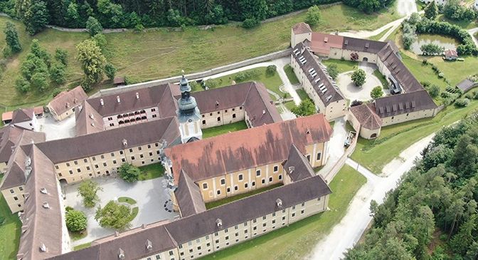 Luftaufnahme eines historischen Schlosses mit mehreren Gebäuden und einem zentralen Turm, umgeben von üppigem grünem Laub und Gras.