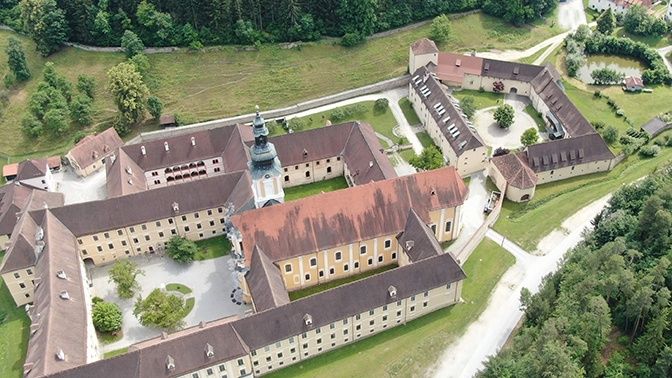 Luftaufnahme eines historischen Schlosses mit mehreren Gebäuden und einem zentralen Turm, umgeben von üppigem grünem Laub und Gras.