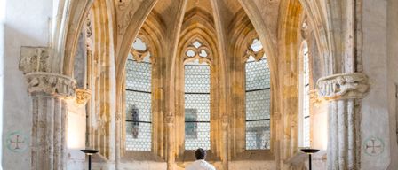 A priest stands before an altar in a chapel with arched windows and intricate stonework.