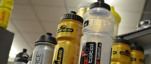 Eine Reihe von Sport-Wasserflaschen, darunter eine transparente mit Bike Total Branding, eine gelbe mit Peeroton Branding und andere, auf einem Regal angeordnet.