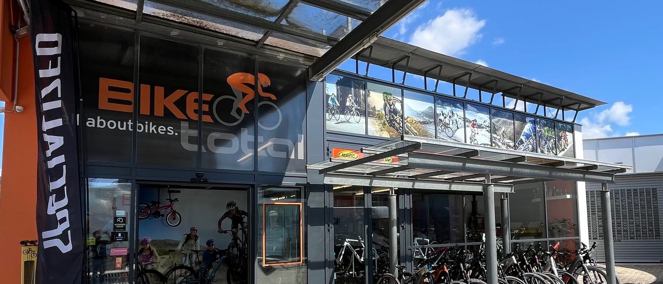 Die Vorderseite eines Fahrradladens namens Bike Total. Es verfügt über große Glasfenster mit Anzeigen und Bildern von Radfahrern. Fahrräder sind draußen vor dem Geschäft geparkt.