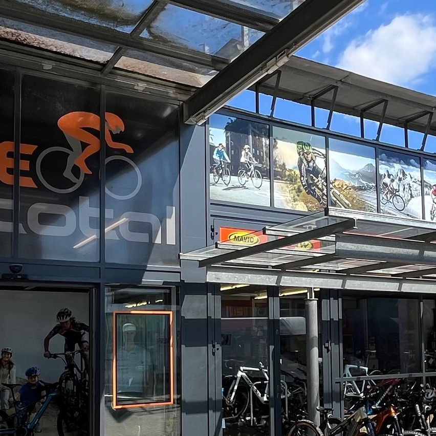 Die Vorderseite eines Fahrradladens namens Bike Total. Es verfügt über große Glasfenster mit Anzeigen und Bildern von Radfahrern. Fahrräder sind draußen vor dem Geschäft geparkt.