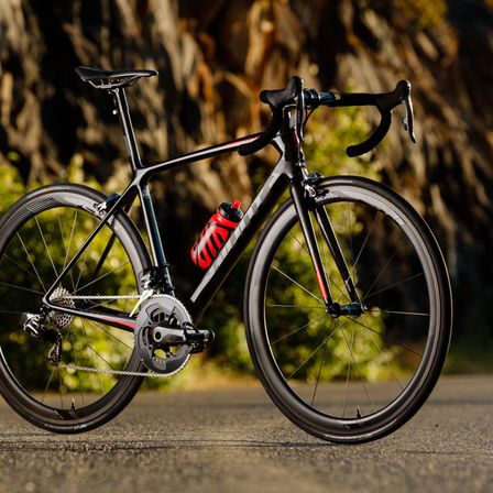 Ein schwarzes Rennrad mit roten Akzenten steht auf der Straße, eine rote und weiße Wasserflasche ist am Rahmen befestigt.