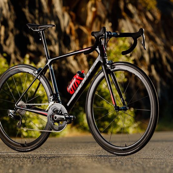Ein schwarzes Rennrad mit roten Akzenten steht auf der Straße, eine rote und weiße Wasserflasche ist am Rahmen befestigt.