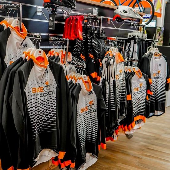 Ein Geschäft, das eine Sammlung von Radsporttrikots zeigt, jedes mit dem Biketoco-Logo. Die Trikots sind in Schwarz, Weiß und Orange. Helme und Radhosen sind ebenfalls an den Gestellen ausgestellt.