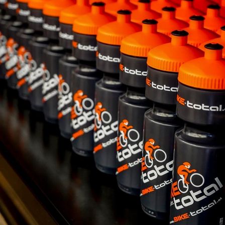 Eine Reihe schwarzer und orangefarbener Wasserflaschen mit dem Text 'BIKE-total' und einem Grafik von einem Radfahrer, die auf einer dunklen Oberfläche aufgereiht sind.