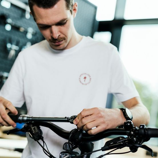 Ein Mann in einem weißen T-Shirt arbeitet an einem Fahrrad und justiert die Lenker. Er befindet sich in einem Laden mit einer Wand voller Werkzeuge und einem Glasfenster.
