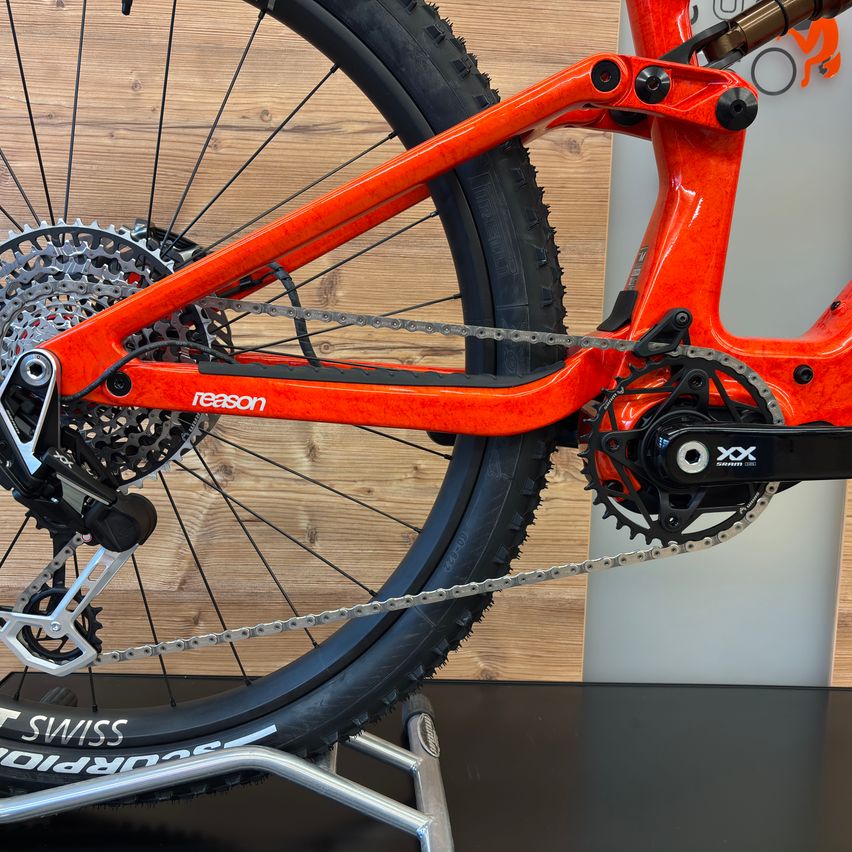 A close-up view of an orange mountain bike with the word 'reason' on the frame. It has a black chain and a black tire. The bike is on a black surface.