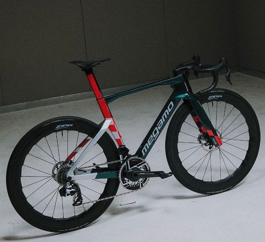 Ein schlankes, schwarzes und türkisfarbenes Rennrad mit roten Akzenten wird auf einem weißen Boden ausgestellt. Das Fahrrad hat einen schwarzen Sattel und Lenker, und das Wort 'megamo' ist auf dem Rahmen geschrieben.