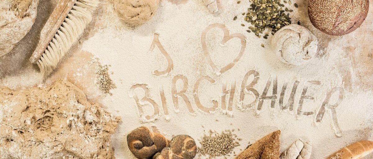 Brot, Samen und Mehl bilden ein Herz und buchstabieren 'Ich liebe Birchermüesli'. Verschiedene Brotsorten, Teigkugeln und ein Pinsel auf einer Holzoberfläche.