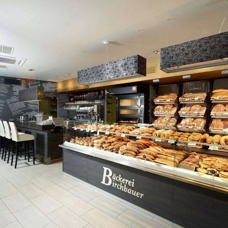 Ein Bäckereiladen mit modernem Design zeigt verschiedene Brot- und Gebäcksorten in einer Glasvitrine. Der Laden hat eine schwarze Wand mit Schriftzug, eine Theke und mehrere Stühle.