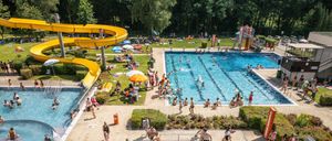 Eine Luftaufnahme eines überfüllten Wasserparks mit mehreren Pools, einer gelben Wasserrutsche und Menschen, die den Tag unter Schirmen und um Bäume herum genießen.