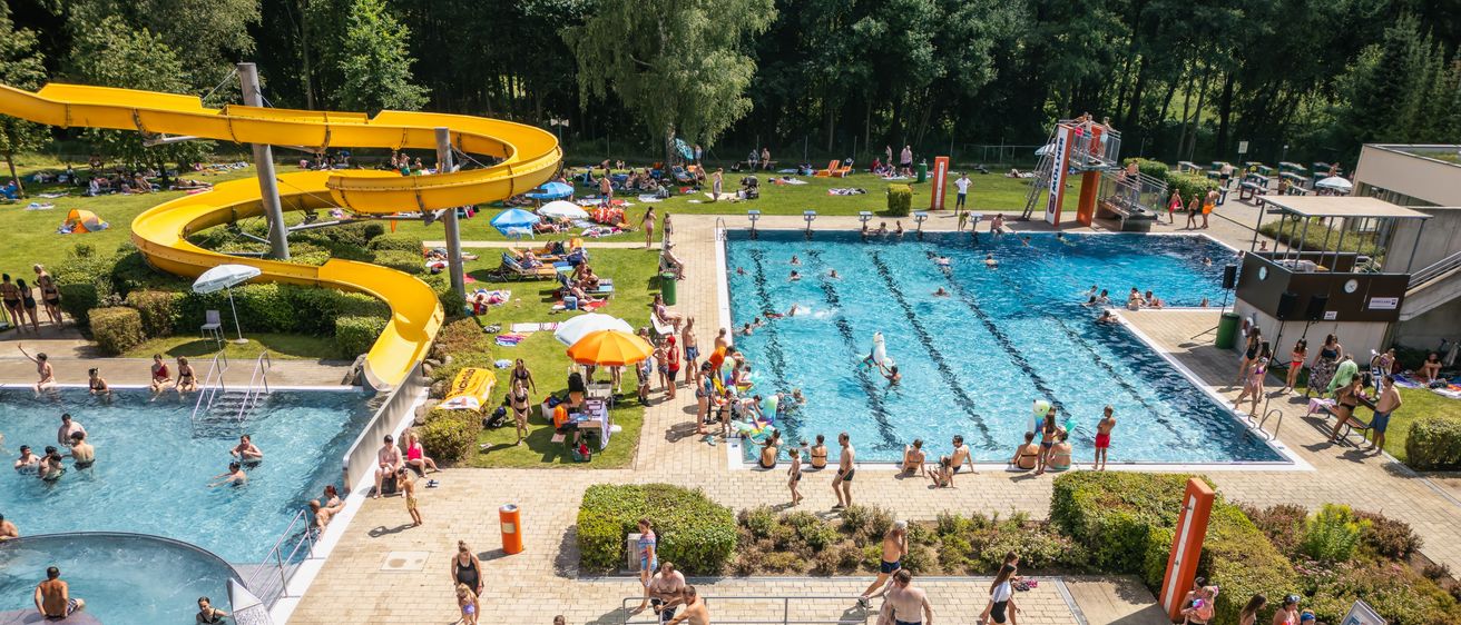 Eine Luftaufnahme eines überfüllten Wasserparks mit mehreren Pools, einer gelben Wasserrutsche und Menschen, die den Tag unter Schirmen und um Bäume herum genießen.
