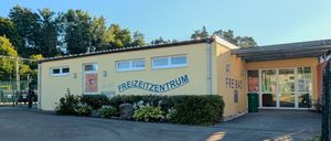 Ein hellgelbes Gebäude mit blauem Text, der Freizeitzentrum Freibad lautet. Es hat Glastüren und Fenster. Rechts steht ein Mülleimer.