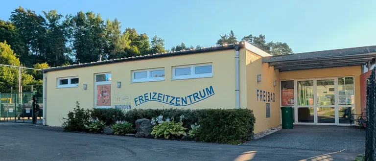 Ein hellgelbes Gebäude mit blauem Text, der Freizeitzentrum Freibad lautet. Es hat Glastüren und Fenster. Rechts steht ein Mülleimer.
