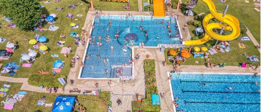 Eine Luftaufnahme eines großen Außenpoolbereichs mit vielen Menschen, die schwimmen, sonnenbaden und spielen. Es gibt zwei Pools, einer mit einer gelben Wasserrutsche. Der Bereich ist von Gras umgeben und hat mehrere Sonnenschirme und Liegestühle. Der Text am unteren Rand lautet 'Letzter Badetag: So., 6. September 2026'.