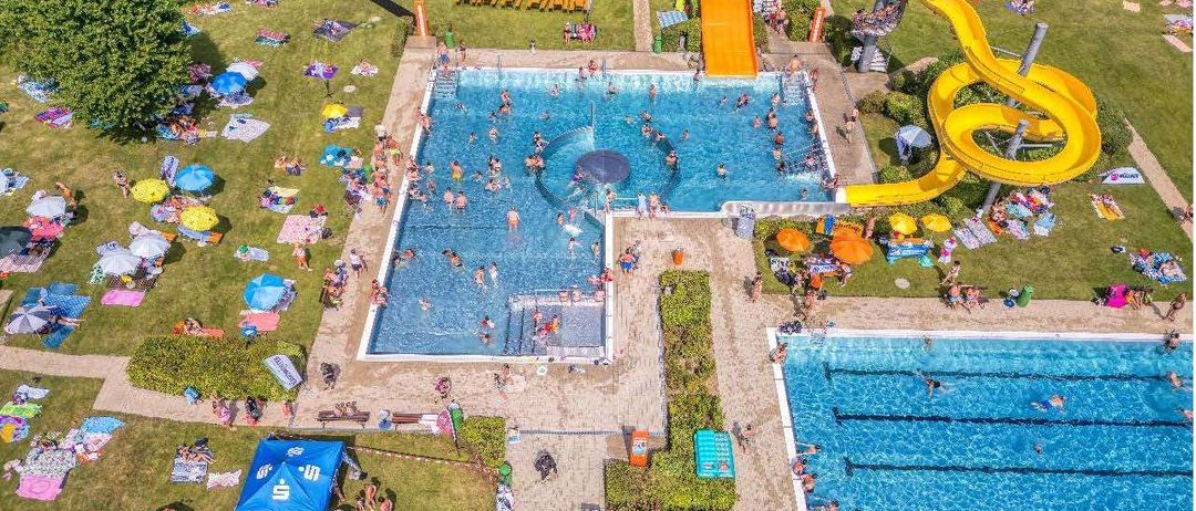 Eine Luftaufnahme eines großen Außenpoolbereichs mit vielen Menschen, die schwimmen, sonnenbaden und spielen. Es gibt zwei Pools, einer mit einer gelben Wasserrutsche. Der Bereich ist von Gras umgeben und hat mehrere Sonnenschirme und Liegestühle. Der Text am unteren Rand lautet 'Letzter Badetag: So., 6. September 2026'.