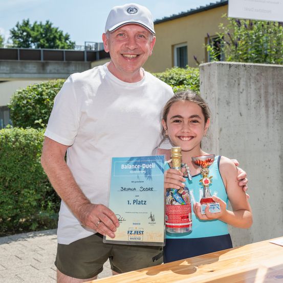 Ein Mann und ein Mädchen halten einen Pokal und eine Auszeichnungsurkunde und lächeln für ein Foto. Der Mann trägt ein weißes T-Shirt und eine Kappe, während das Mädchen ein blaues Tanktop trägt. Sie stehen neben einem Holztisch mit einer Flasche und einem Becher darauf. Dahinter steht ein Gebäude mit einem Balkon und etwas Grün.