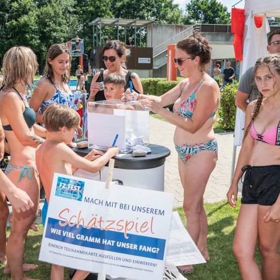 Eine Gruppe junger Menschen und Erwachsene versammelt sich im Freien. Zwei Frauen helfen Kindern, Steine in eine Box zu legen. Die Box hat ein Schild mit der Aufschrift 'Schatzspiel: Wie viel Gramm wiegt unser Schatz?' Die Veranstaltung scheint sich an einem Strand abzuspielen.