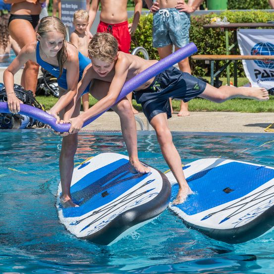 Zwei Kinder stehen auf Surfbrettern im Pool und halten eine lila Röhre. Ein Banner ist hinter ihnen sichtbar.