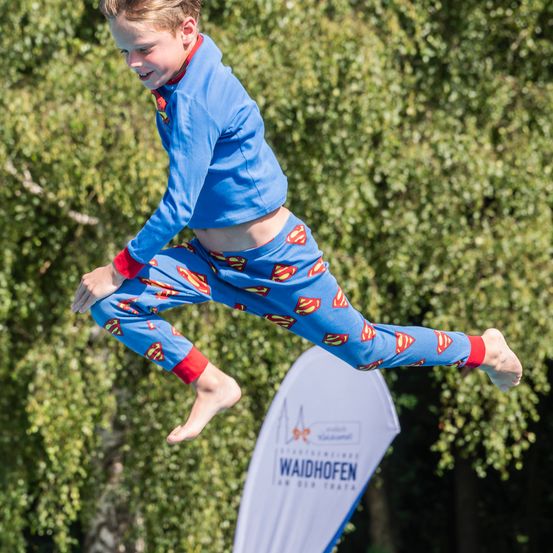 Ein junger Junge in blauen Superman-Pyjamas springt in der Luft über ein weißes Schild, das Waidhofen steht.