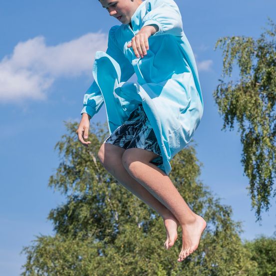 Ein Junge in einem blauen Gewand und Shorts springt mit ausgestreckten Armen in der Luft unter einem blauen Himmel mit Wolken.