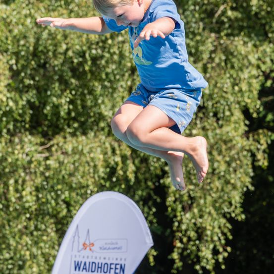 Ein junger Junge springt in die Luft in der Nähe eines grünen Busches, trägt ein blaues Shirt und Shorts. Unten steht ein weißes Schild mit blauem Text, das Waidhofen liest.