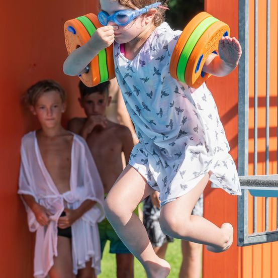 Ein kleines Mädchen springt mit bunten Schwimmhilfen in den Händen, während zwei Jungen hinter ihr stehen und zusehen.